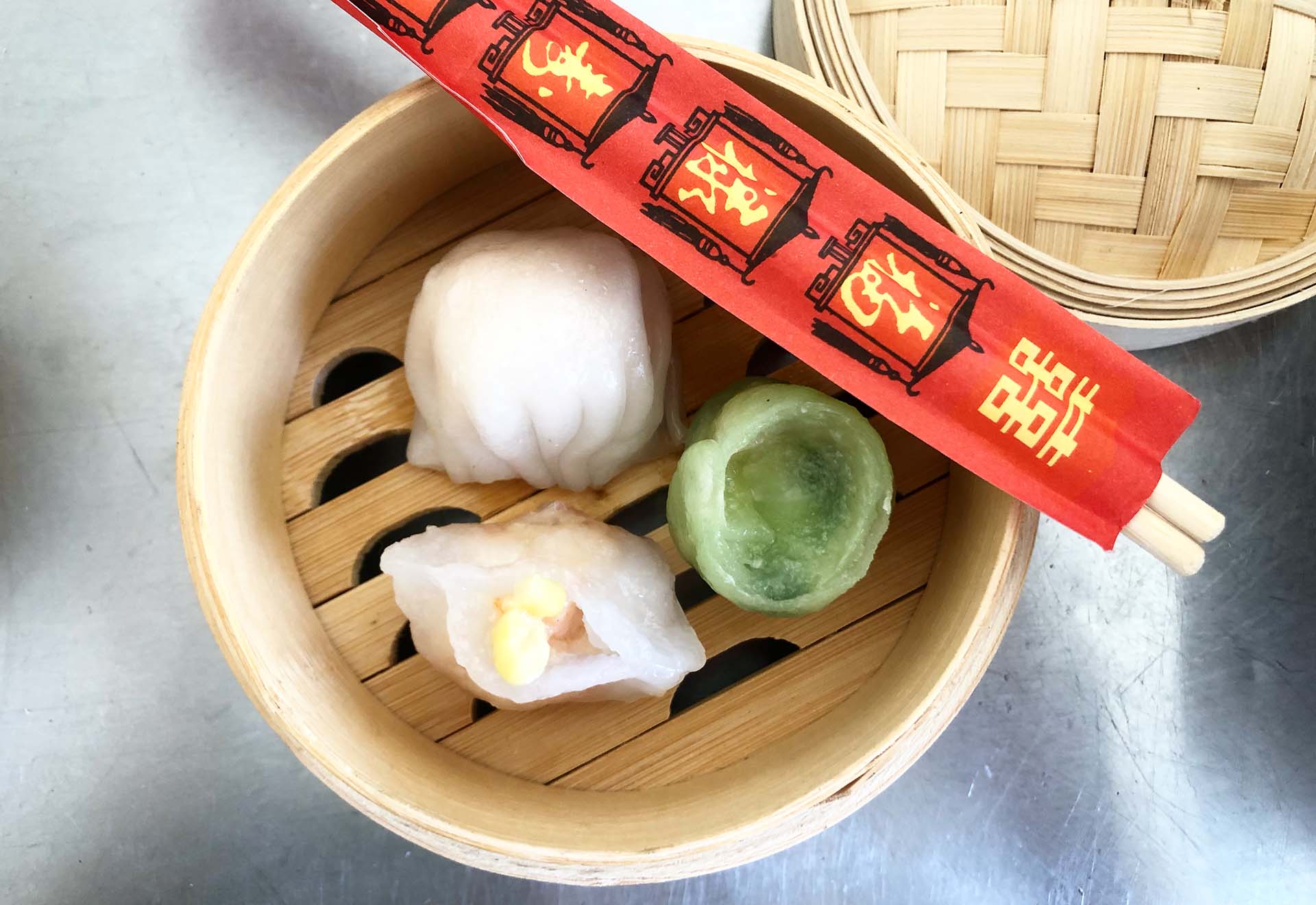 Three different types of dumplings in a bamboo steamer, with a pair of chopsticks in a red paper sleeve resting on top.