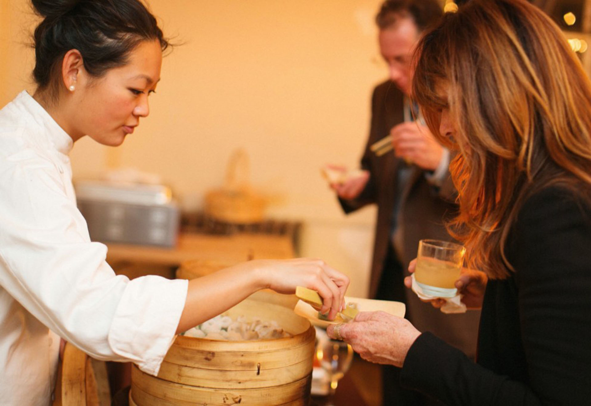 A young woman in a white chef's jacket serves dim sum to a guest at a fancy gala.