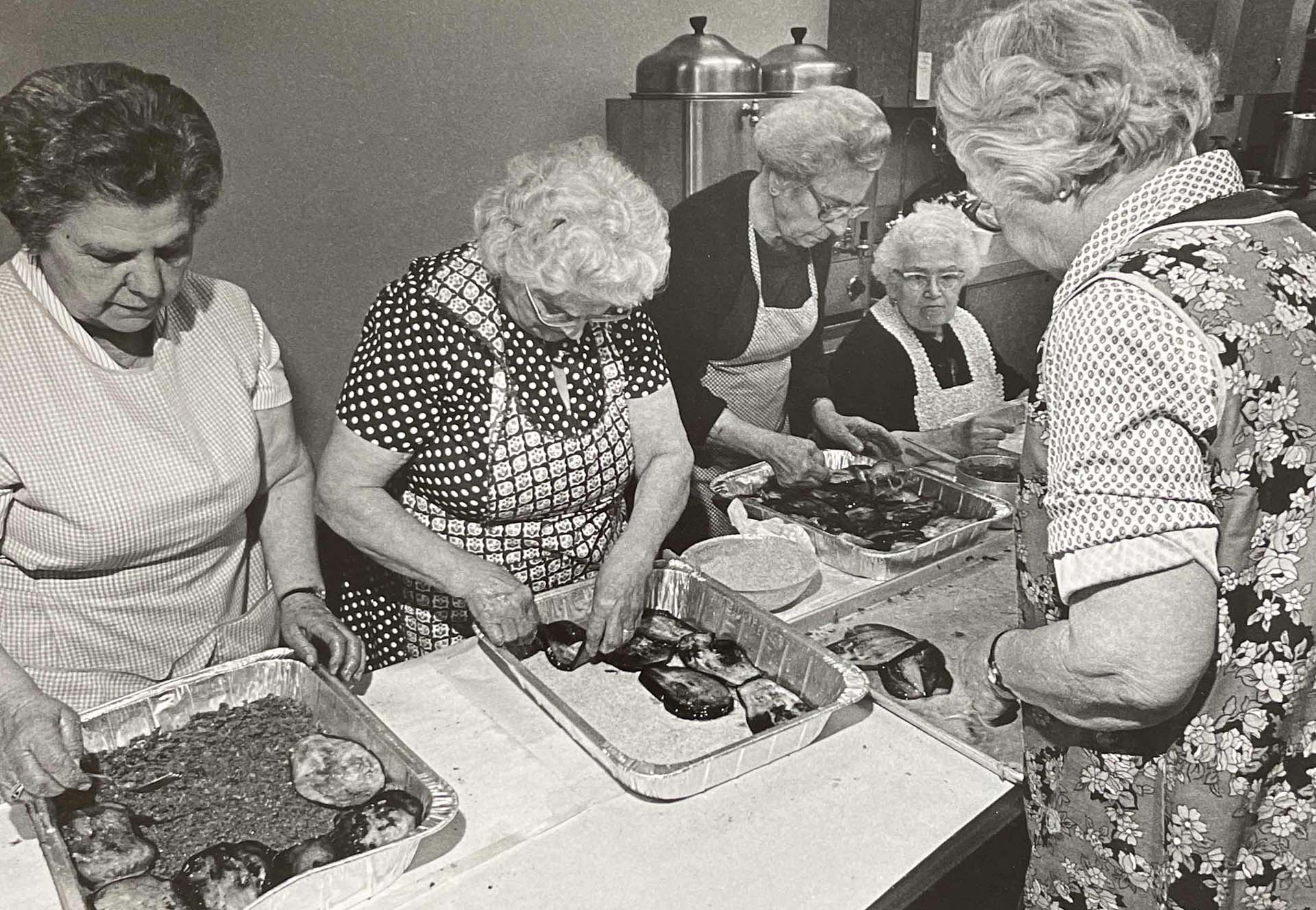 Black and white photo of elderly women in aprons preparing trays of Greek moussaka.