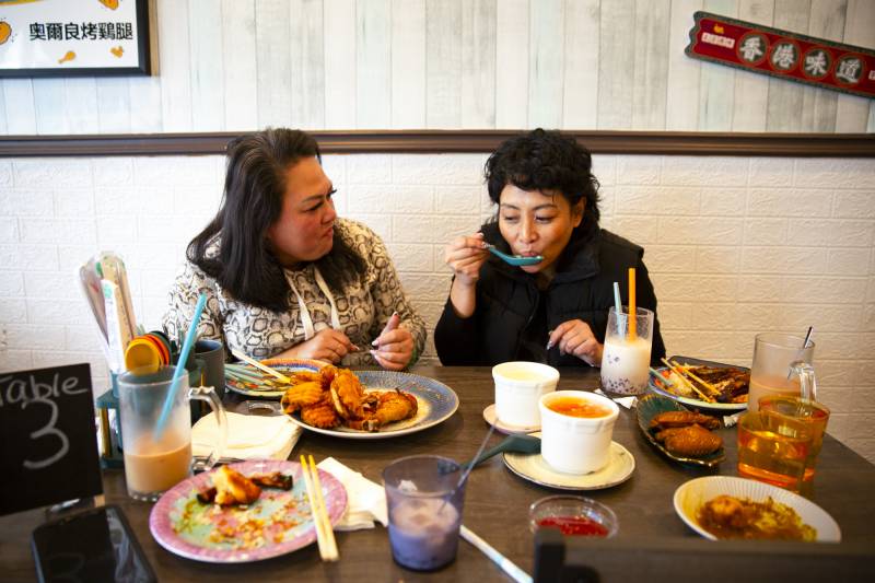 Two women sit at a large round table inside a Chinese restaurant, a spread of chicken wings and other dishes in front of them.