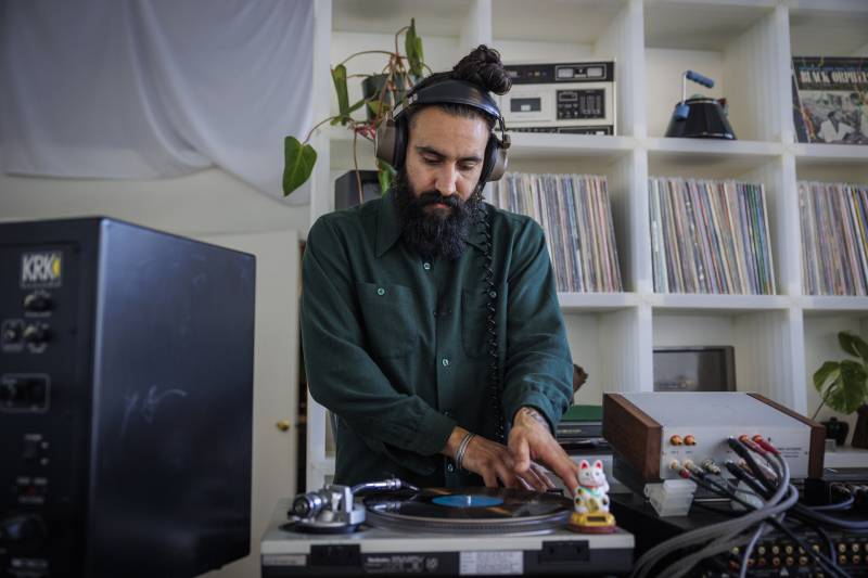 A man with a beard and man-bun stands in front of a record player, operating it.