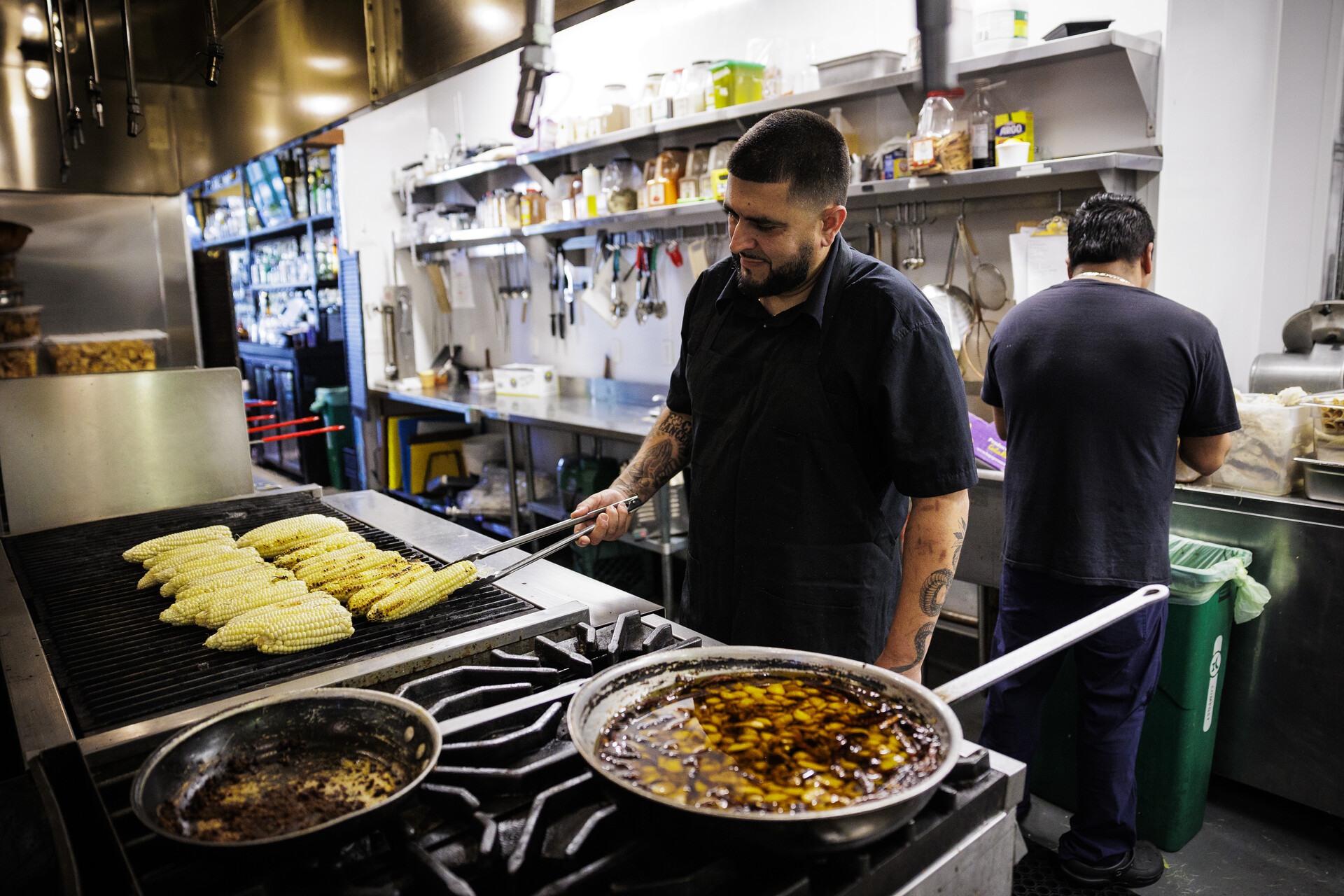 A chef uses tongs to turn ears of corn cooking on the grill inside a restaurant kitchen.
