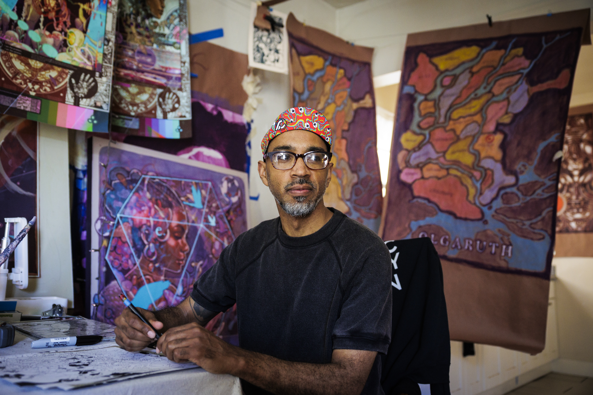 Black man with glasses sits in a room filled with bright paintings and drawings