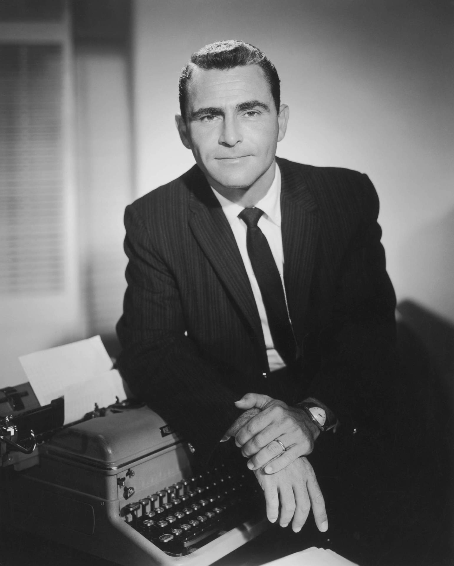 A black and white image of a white man in a 1950s-era suit, leaning on a typewriter.