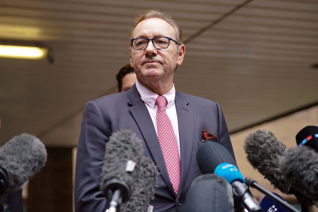 A white man in a grey suit and pink tie stands behind a podium with many microphones at chest height.