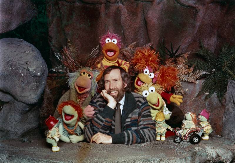 A smiling bearded man wearing a tie and a cardigan sits smiling surrounded by colorful puppets on a soundstage.
