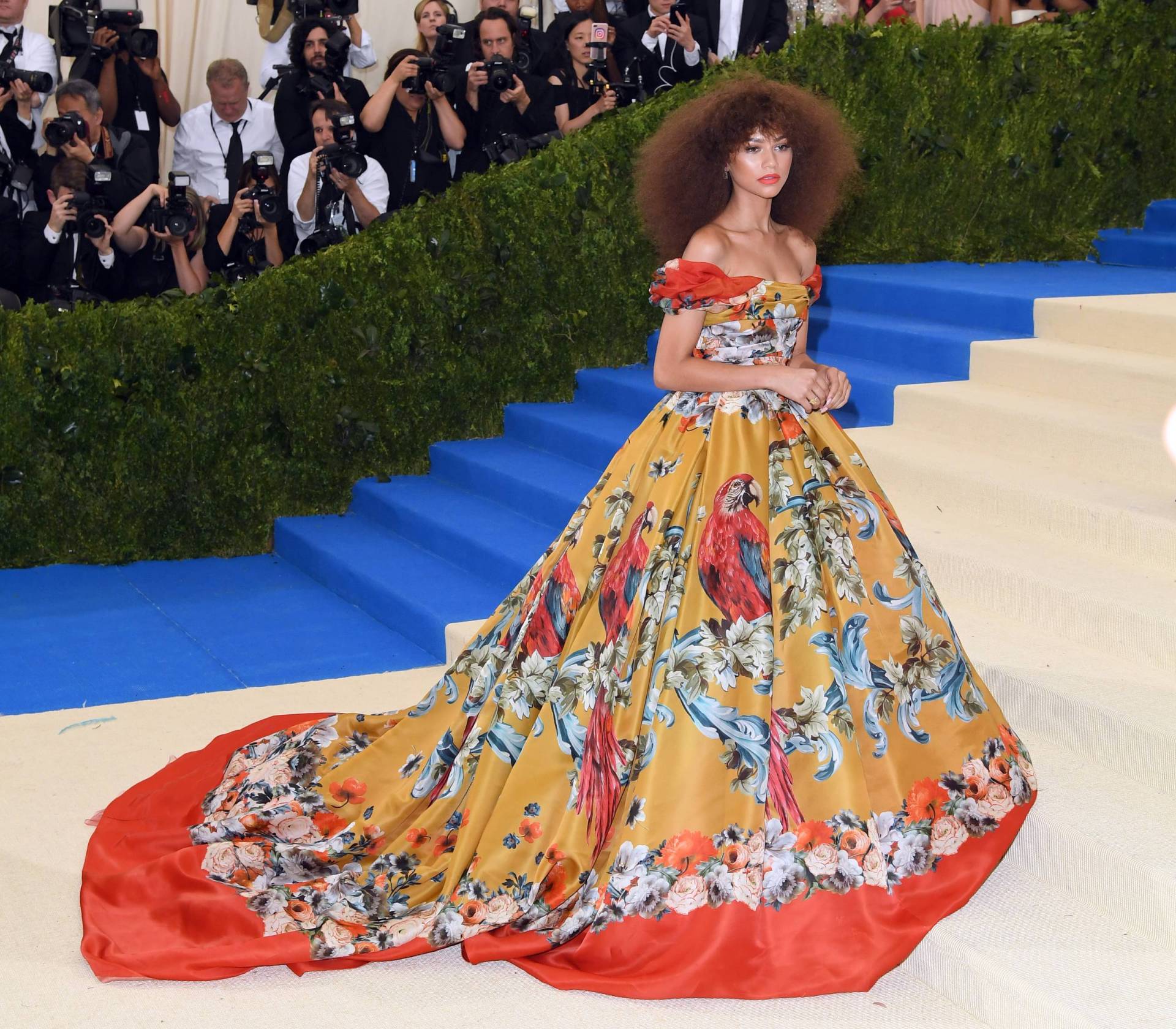 A young Black woman with natural hair stands at the foot of a staircase wearing a red and orange off the shoulder gown featuring a bold pattern including parrots. 