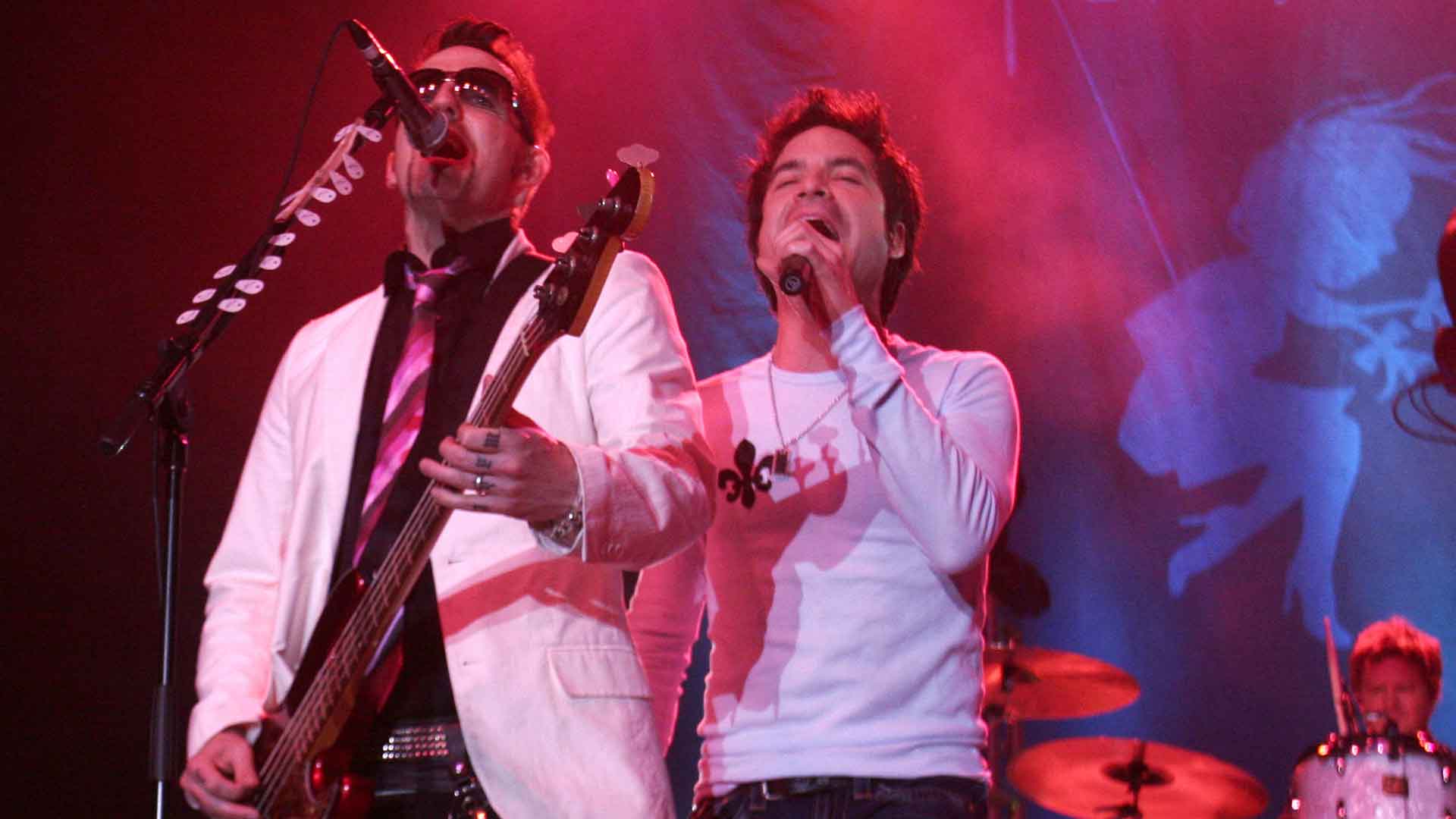 A man in sunglasses plays the bass guitar while a man sings next to him