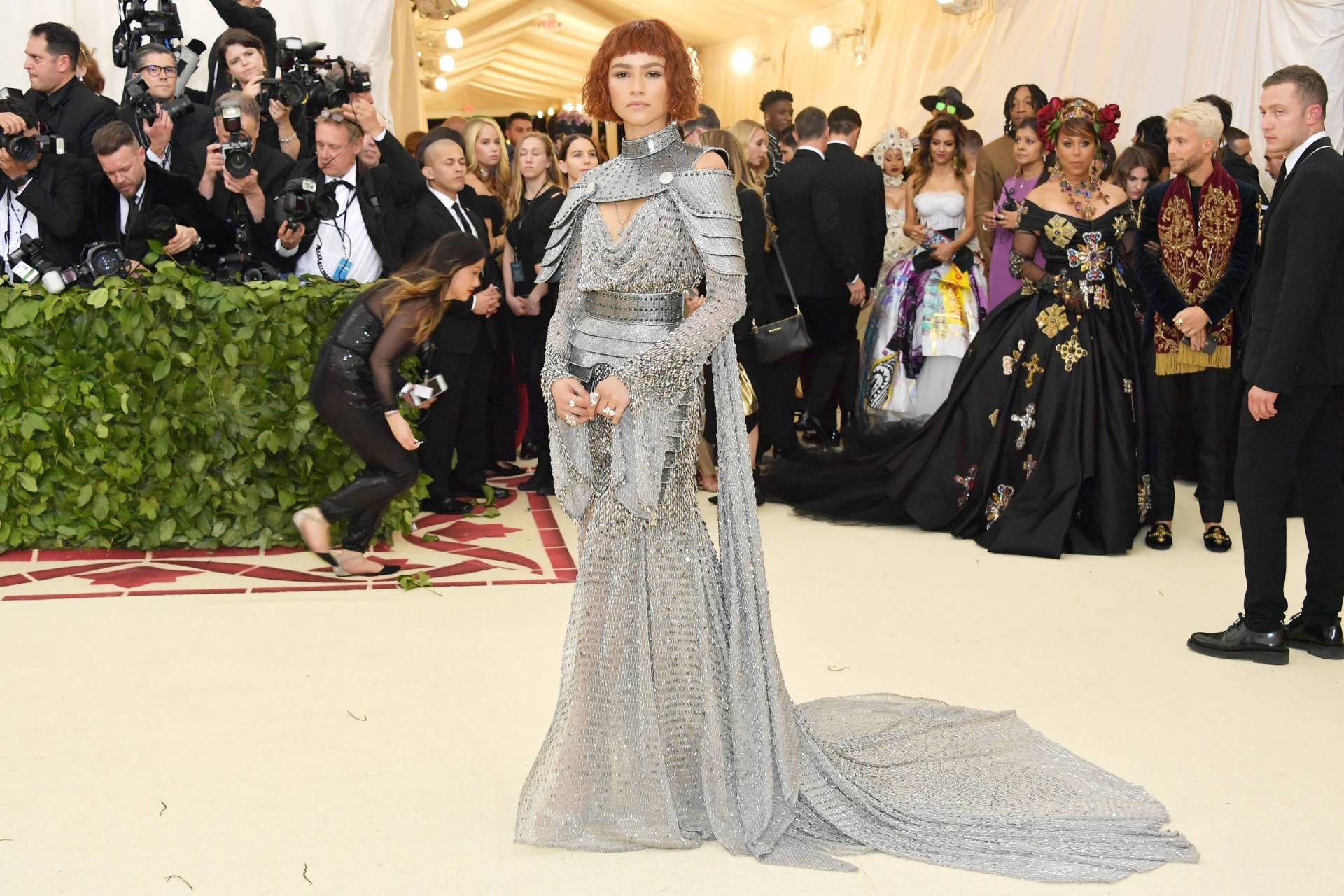 A beautiful young woman with a red bob stands on a white carpet surrounded by photographers, wearing a dress made of chainmail and armor.