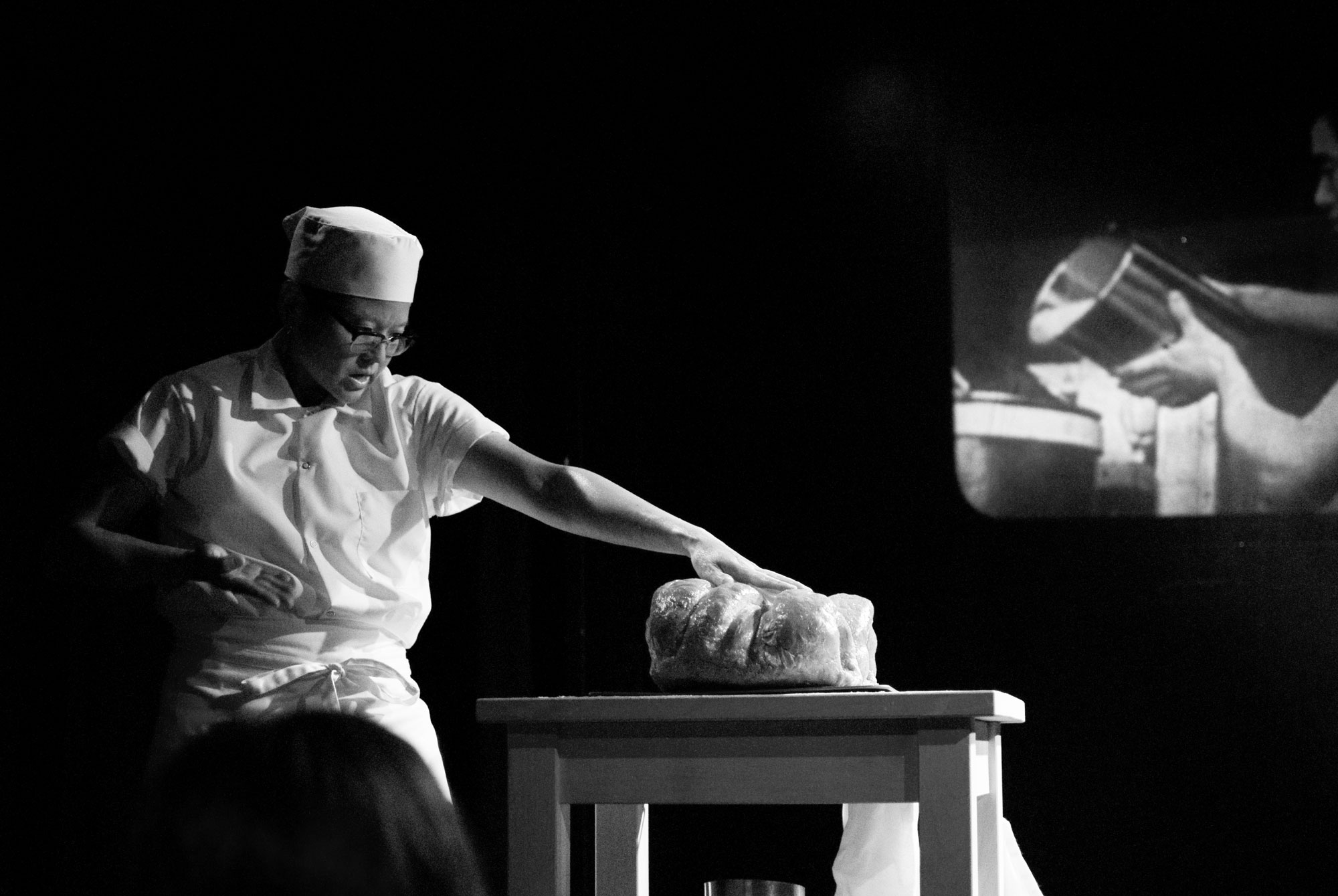 Black and white image of person dressed as baker interacting with a loaf of bread, projection behind