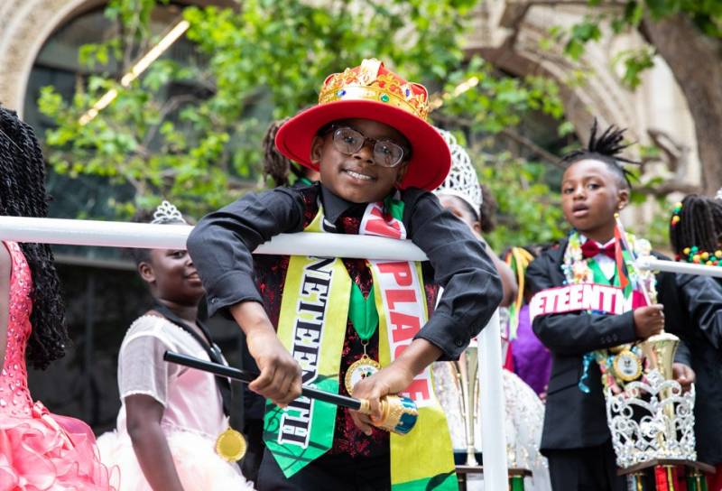 A child with a fancy orange hat, dress clothes and a sash rides on a float during the Juneteenth parade and festival.