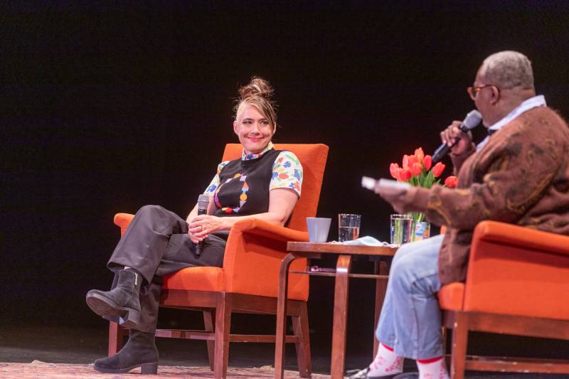 On a theater stage, a middle aged white woman with a messy up-do sits in an orange armchair smiling at a Black man, seated in matching armchair near her. They are separated by a small table with a vase of tulips on.