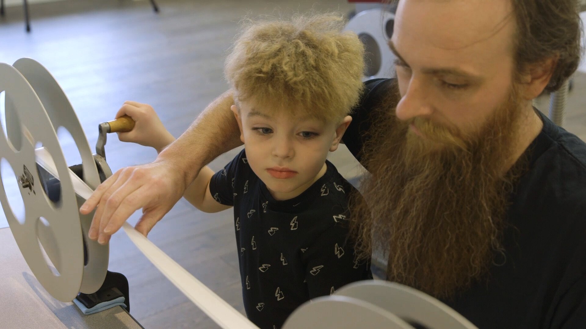 child winds a film reel under instruction of a man with a beard