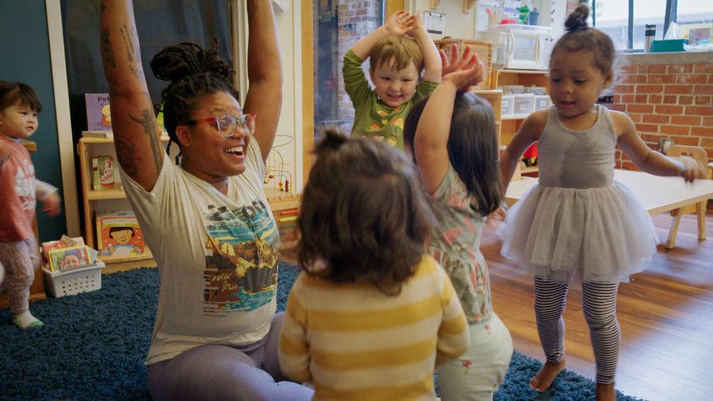 Adult leads a circle of young children in an exercise, their arms raised