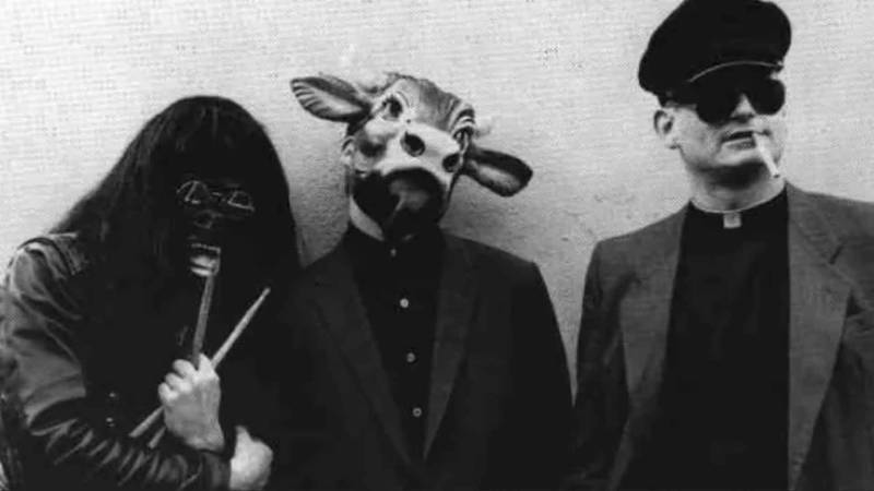 A man in a gasmask holding drumsticks, a man in a cow mask, and a man dressed as a priest, smoking a cigarette