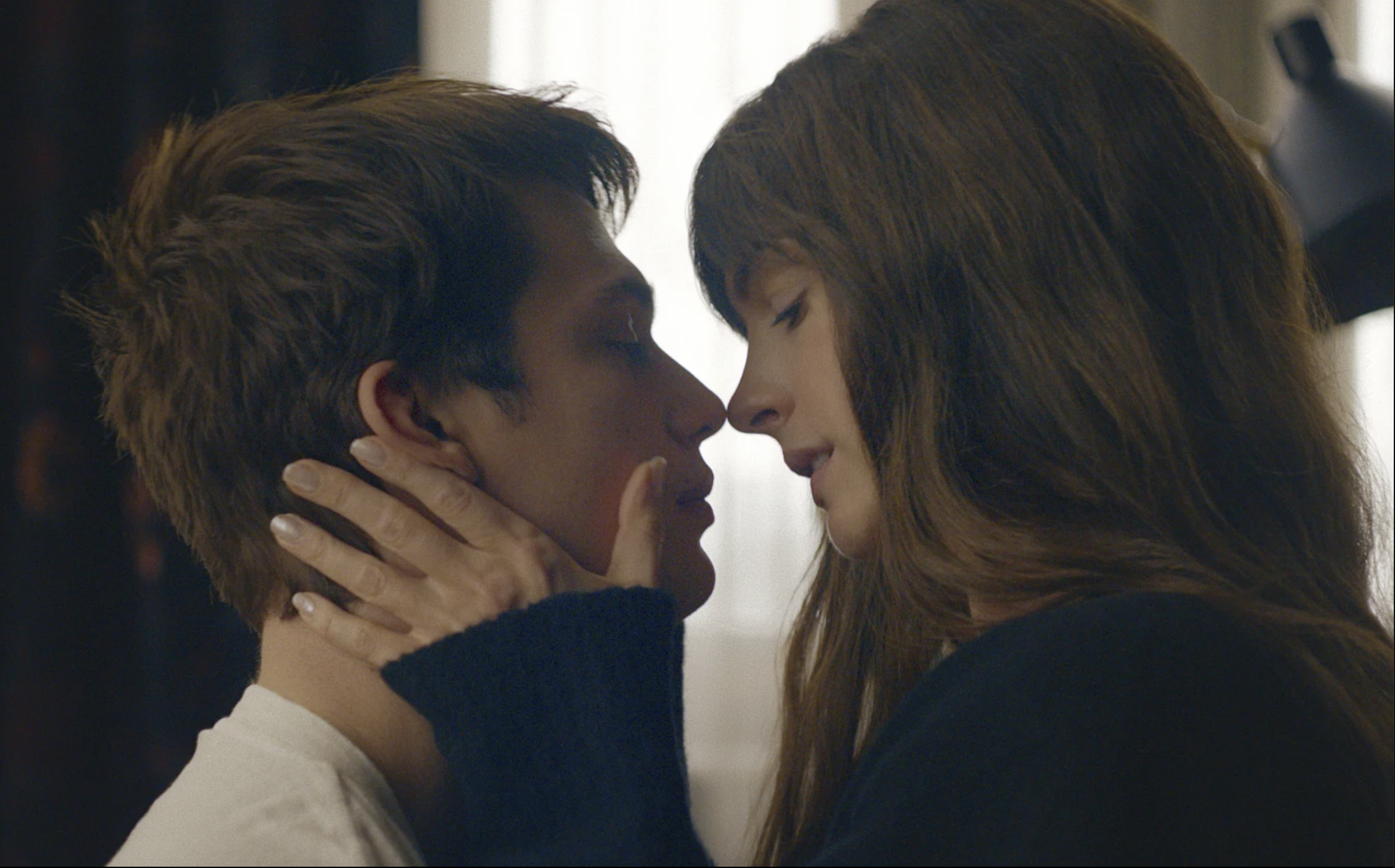 A white woman with long brown hair and younger white man lean in to kiss.