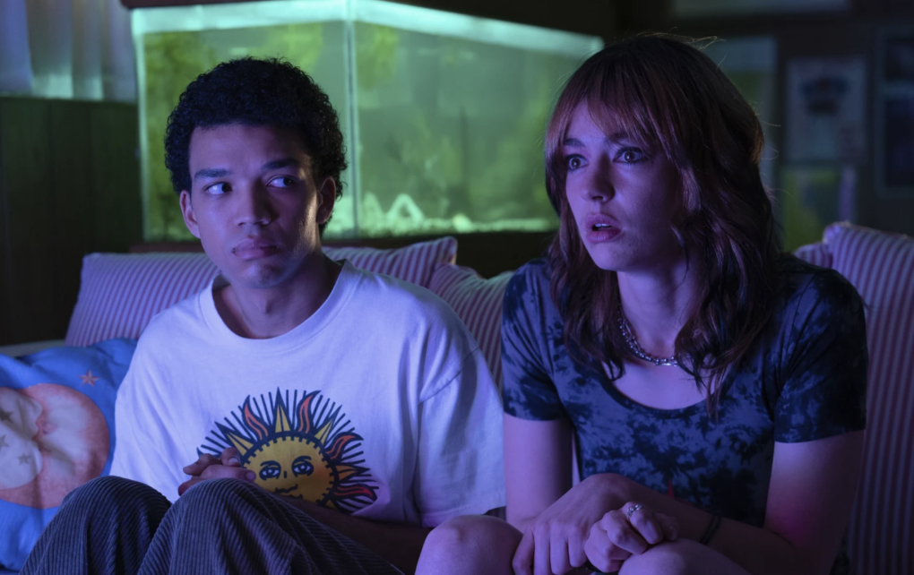 A Black teenage boy and a white teenage girl sit on a couch with a glowing fish tank behind them.