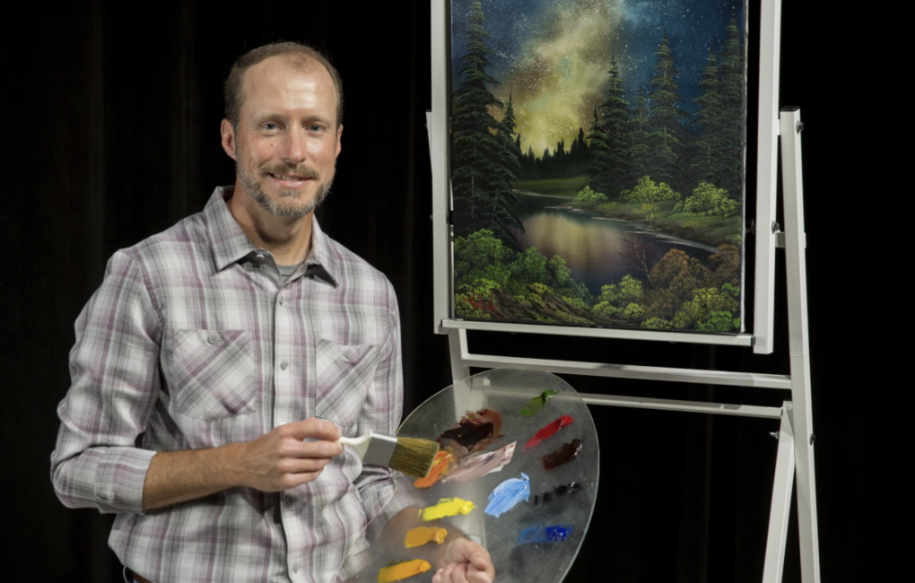 A middle aged man wearing a plaid shirt stands next to a painting. He is holding an easel and a brush.