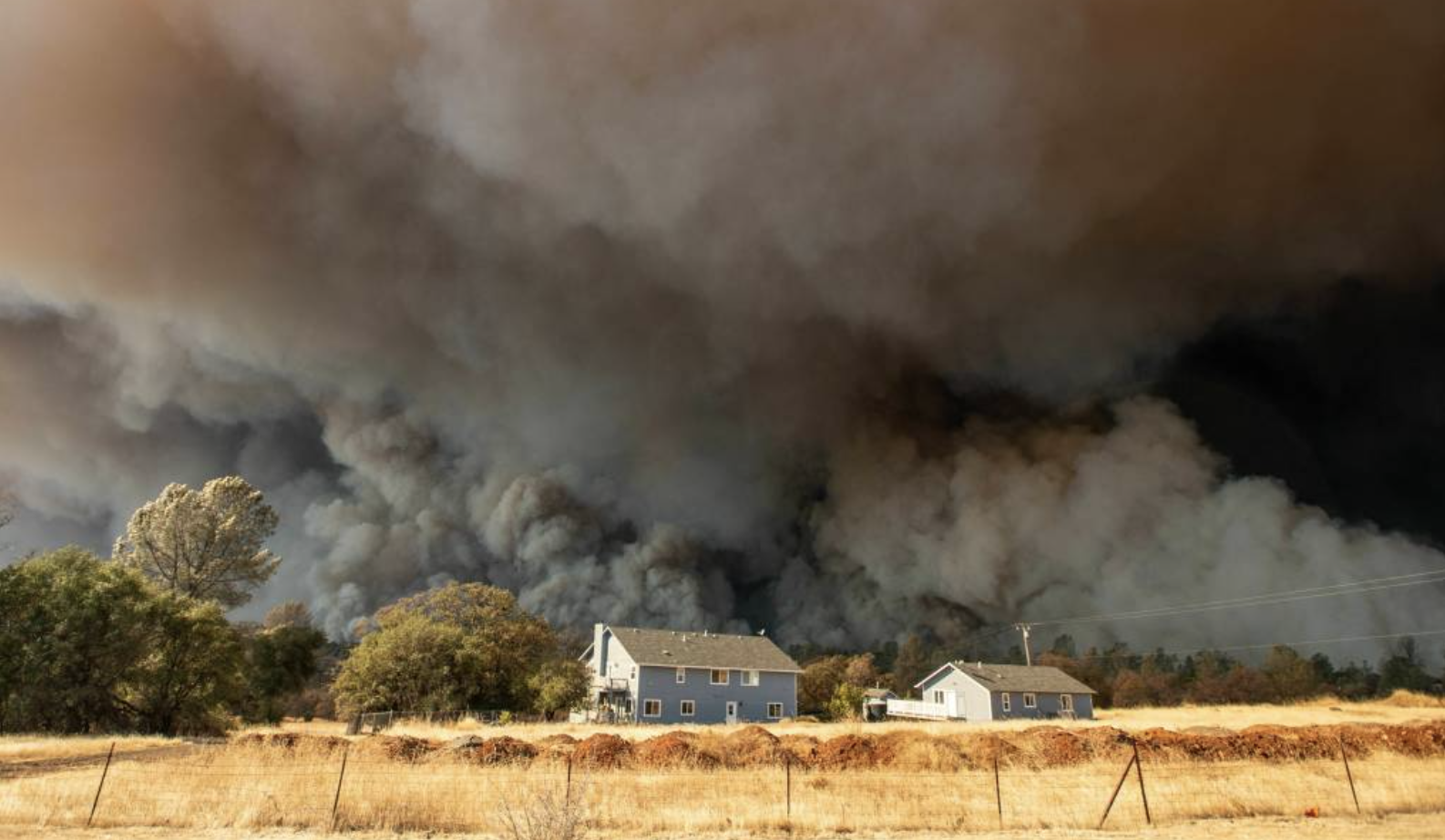 A blue house in a rural environments is surrounded by plumes of Grey and black smoke.