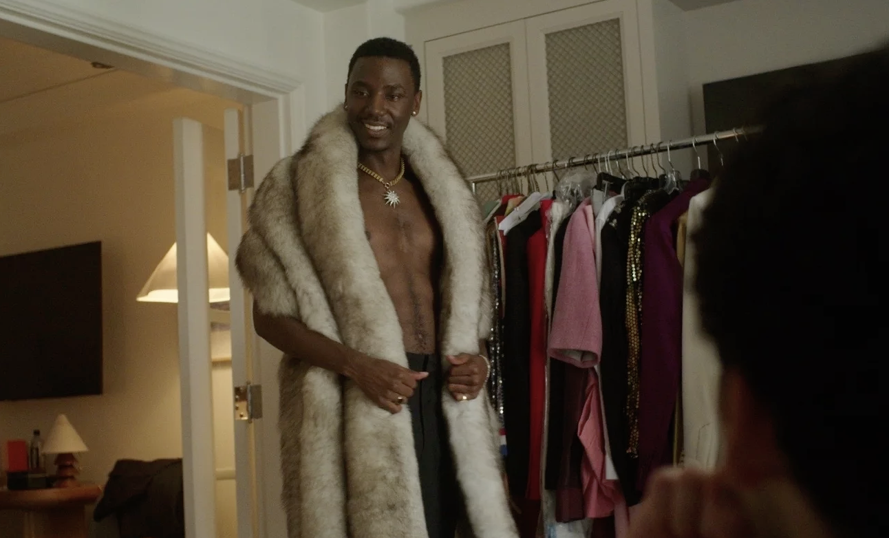 A young Black man stands next to a clothes rack, shirtless and wrapped in a fur stole.
