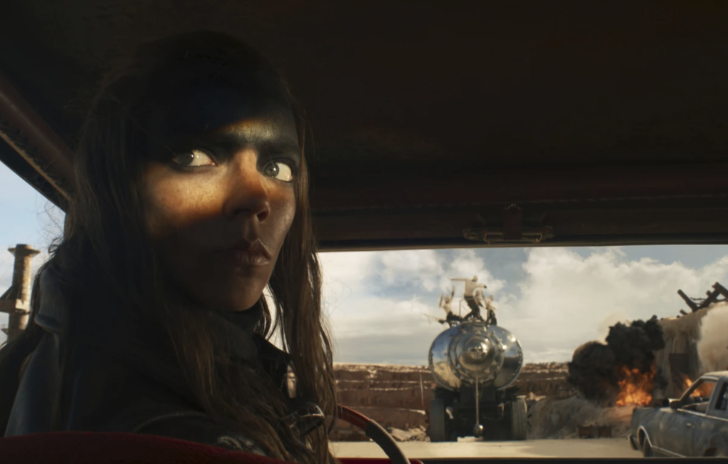 A young woman viewed from behind inside a car, peers over her shoulder from the driver's seat. Through the windscreen, a large truck burns.