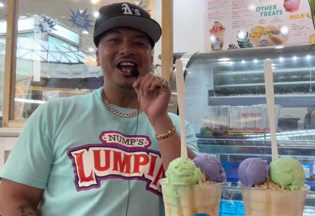 Screenshot from a video: A man in a "Nump's Lumpia" T-Shirt laughs, with two glasses of halo halo in front of him.