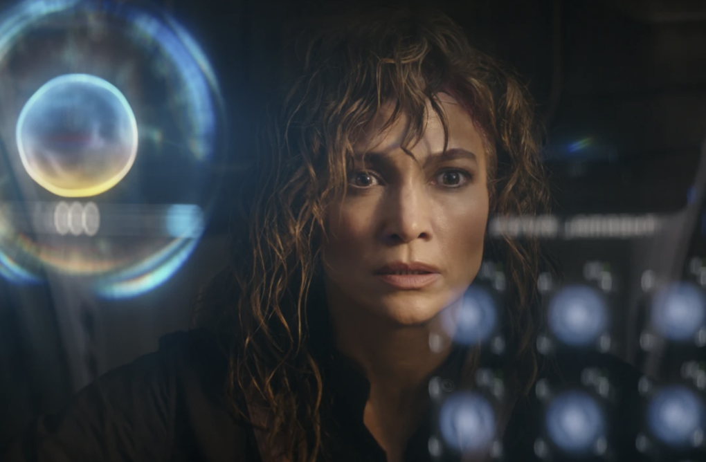 A nervous looking woman with long bedraggled hair stares at a futuristic screen, looking very concerned.