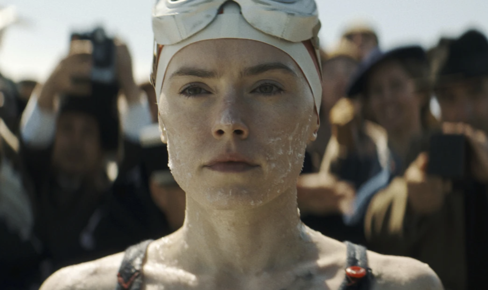 A young woman wearing a swimsuit, a swimming cap and goggles, with grease on her face, stands with a crowd behind her, serious expression on her face.