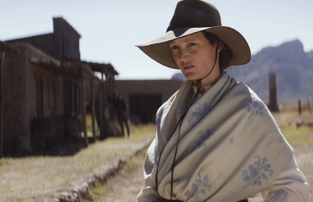 A woman stands in front of old west style wooden buildings. She is wearing a broad rimmed hat and a belted blanket over her clothes.