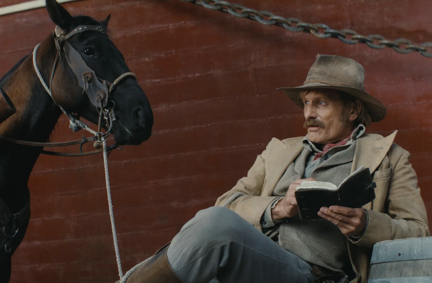 An old west cowboy sits next to his horse on a bench, reading a book.