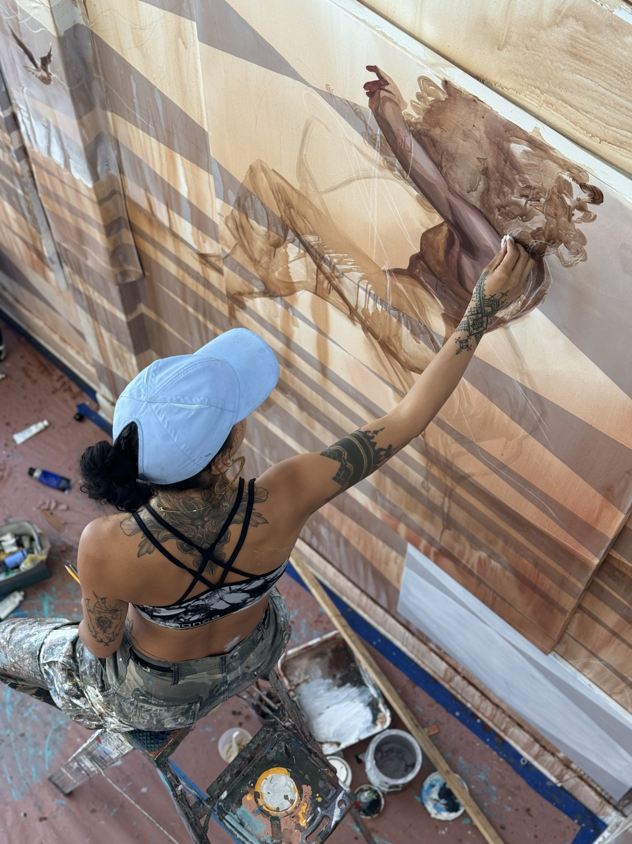 Woman with blue hat on paints a brown and gold image on a canvas.