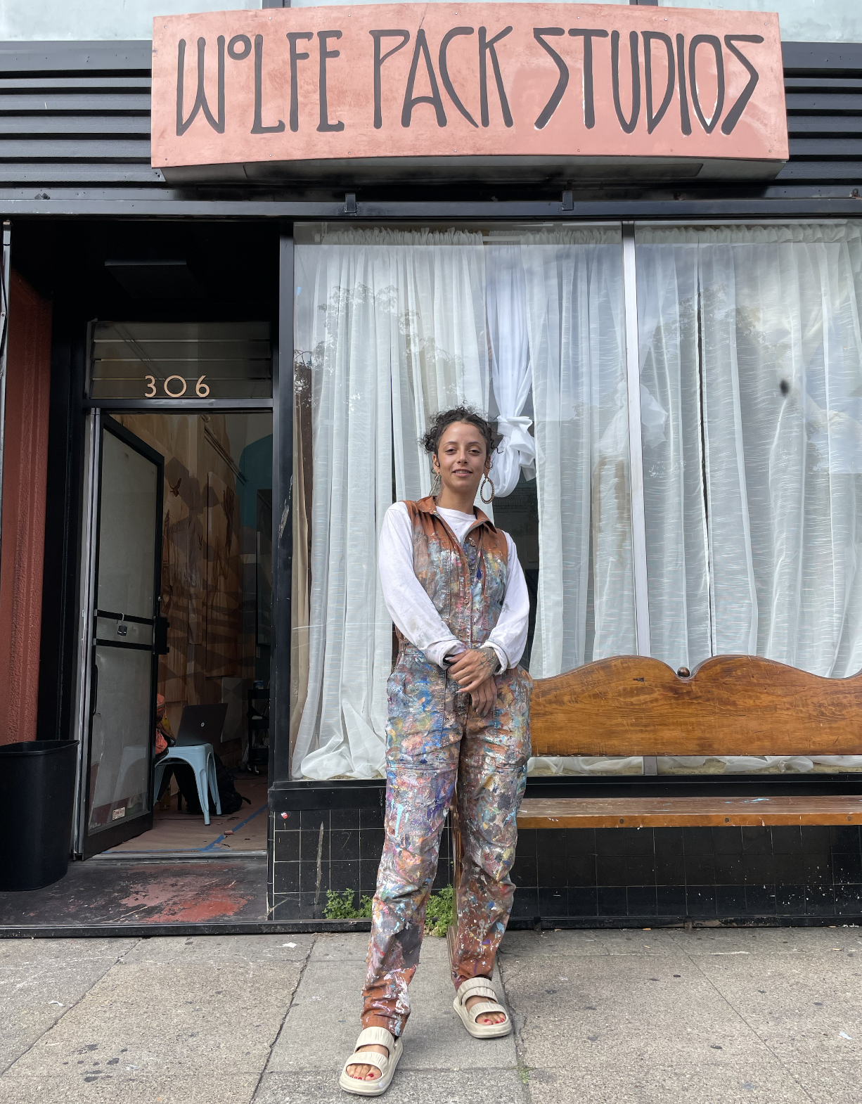 Woman wearing clothes covered in different colors of paint standing in front of a studio-gallery space.