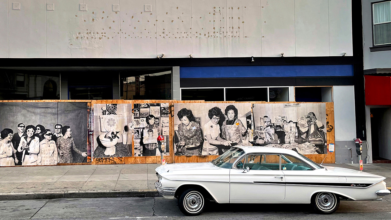 Classic car in front of wall of wheatpasted black and white family photos on boarded-up storefront