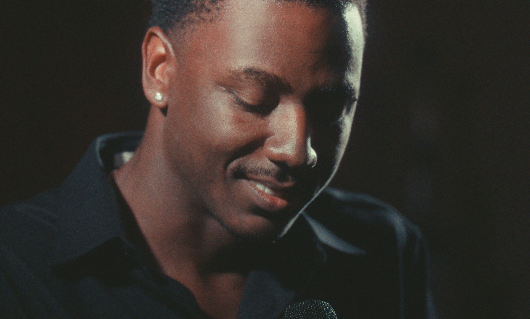 Close up on a young Black man's face and shoulders. He is glancing downwards and smiling.