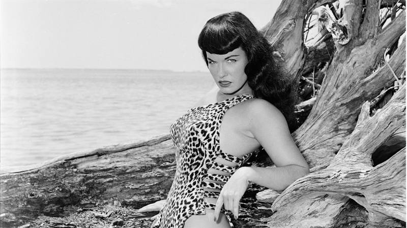A beautiful white woman in the 1950s, wearing a leopard print one-piece swimsuit and a fierce facial expression, leans against a rock formation at the beach.