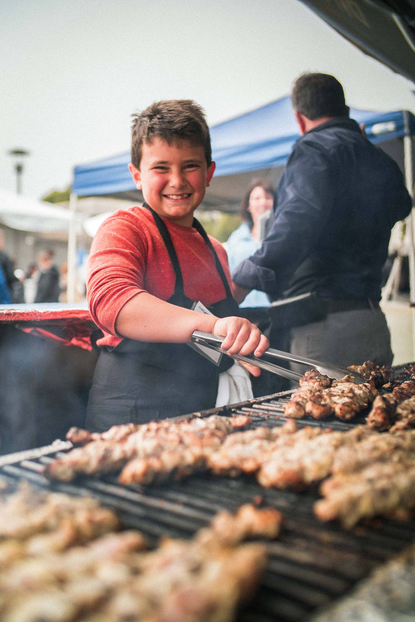 A smiling young boy holds a pair of metal tongs as he tends to lamb kebabs cooking on the grill.
