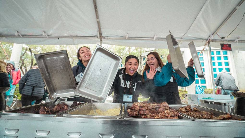Three young women hold up the lids from a steam table, revealing large piles of grilled lamb.