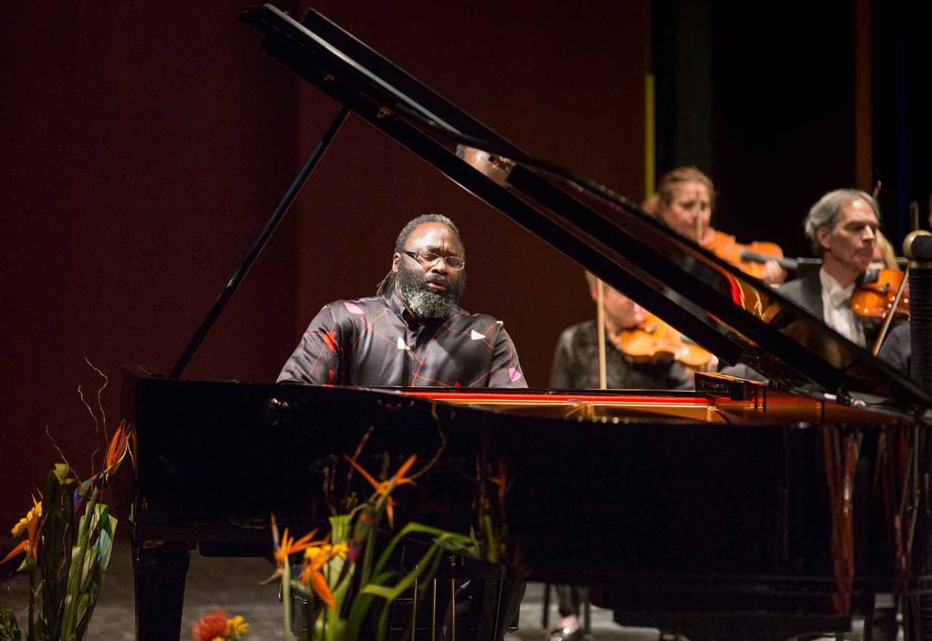 A Black man with a beard in a black and red jacket plays a grand piano.