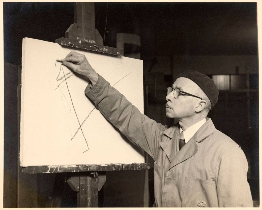 Black and white photo of man in glasses drawing lines on a canvas