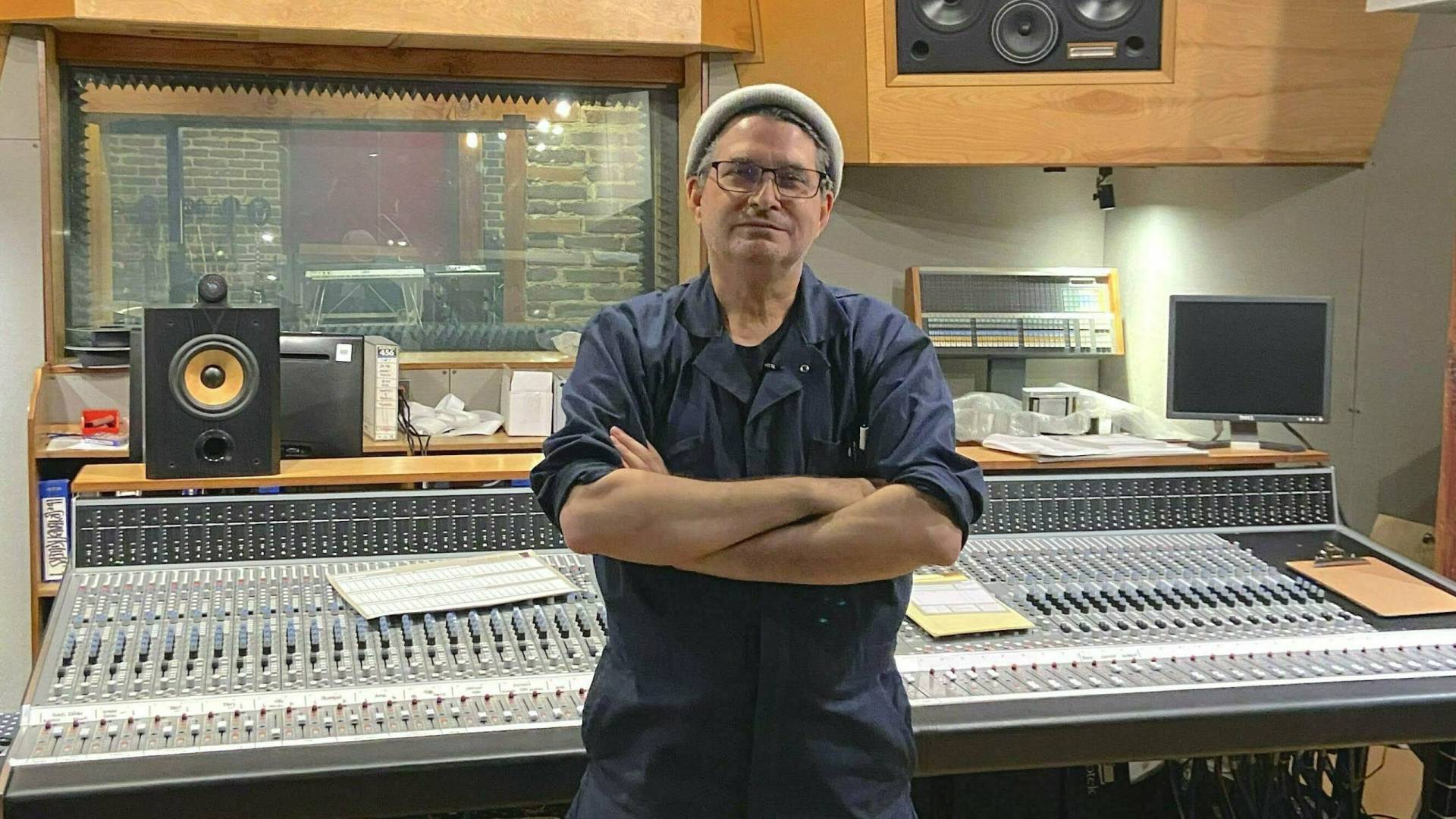 A man in overalls and a beanie stands, arms folded, in front of a music studio control desk.