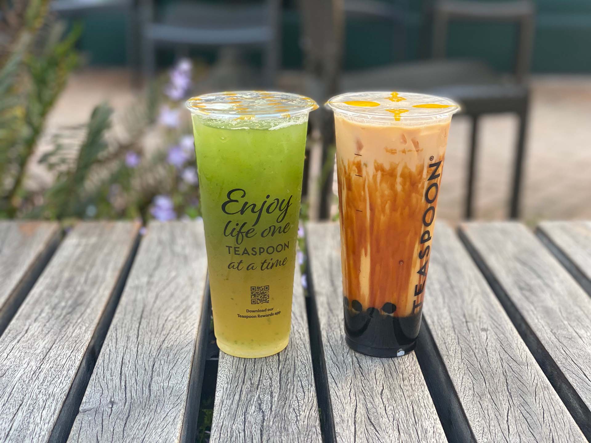 Two boba drinks — one green and fruity, the other one creamy — on a wooden picnic table.