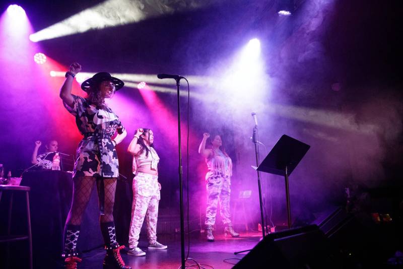 Women on a smokey stage with purple lighting holding their fist up. 