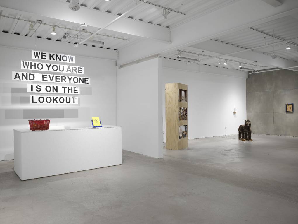 White-walled space with hanging text piece over desk, video tower, sculpture and wall work
