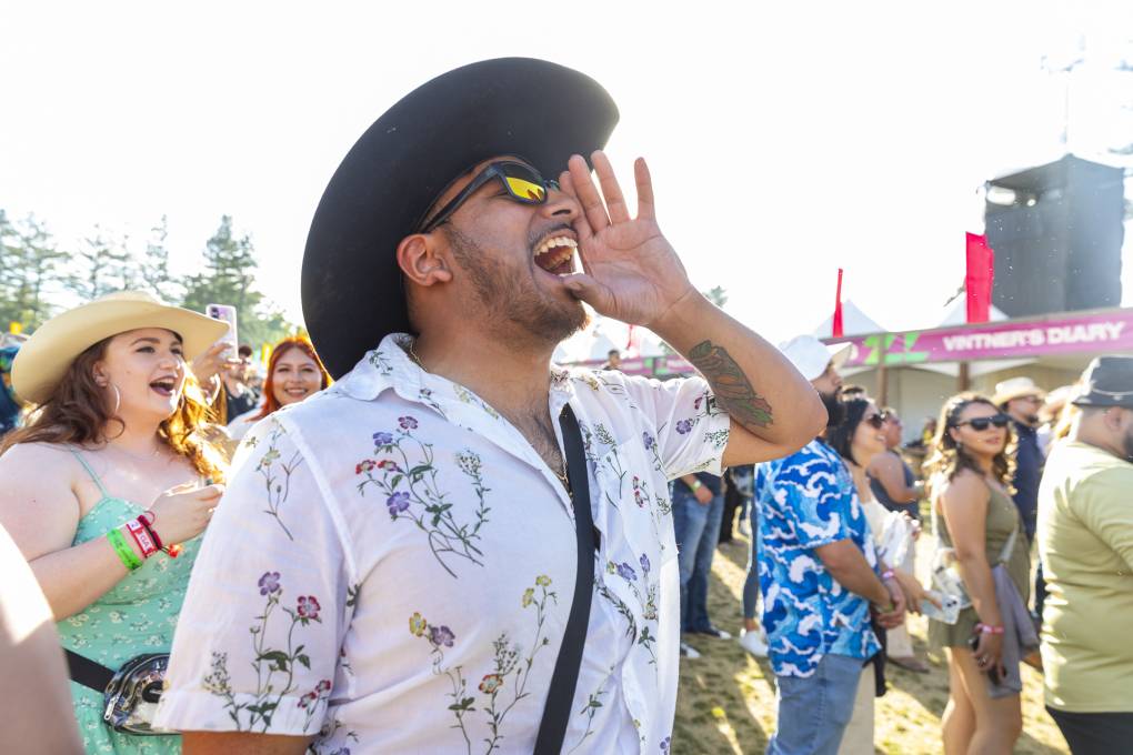 A man in a white button-up shirt and large black cowboy hat holds up his left hand to his mouth, shouting to performers