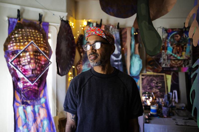 A Black man wearing glasses stands in an art studio, looking to his right