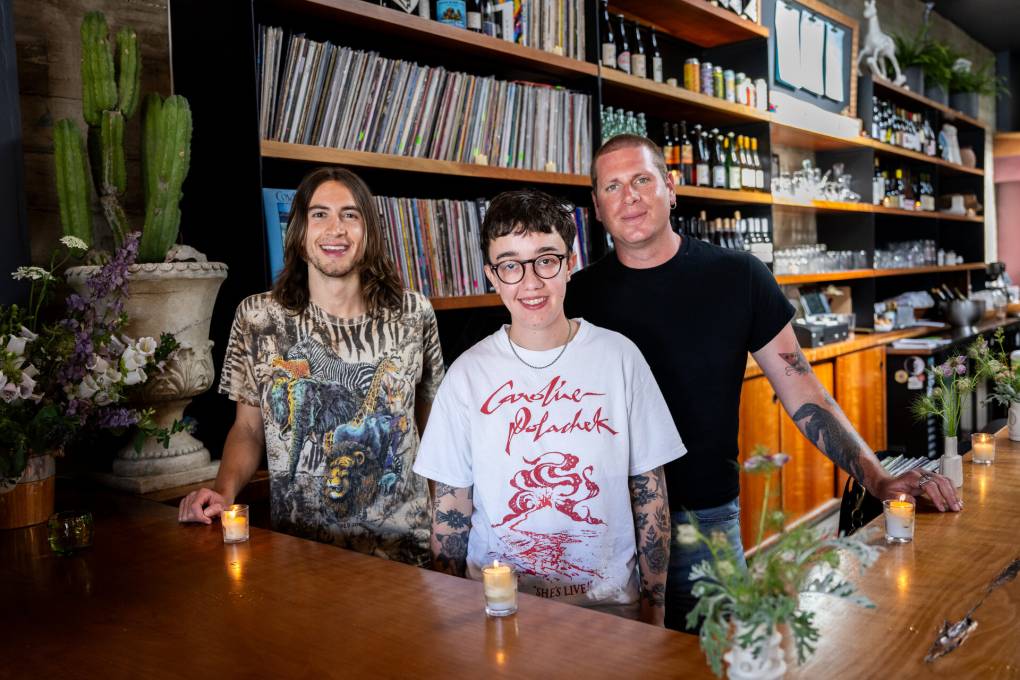 Three people in T-shirts stand behind a service counter, with a wall of vinyl LPs in the background.