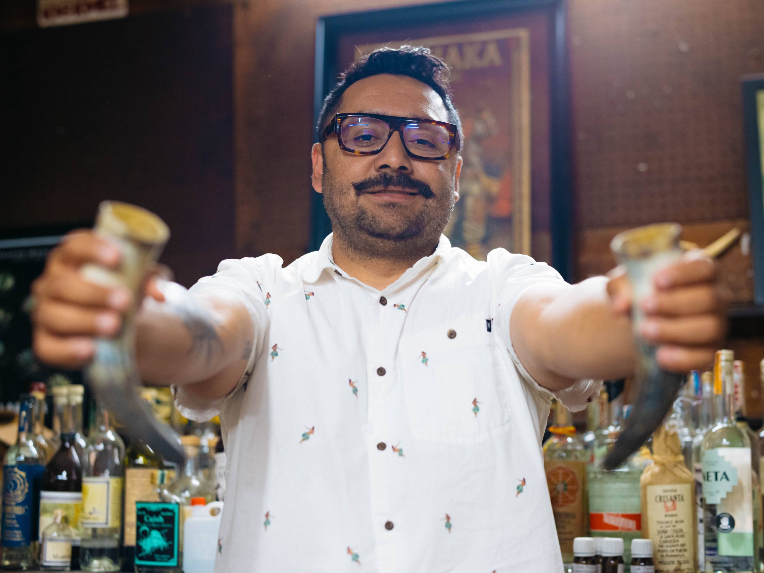 a man holds up two shots of mezcal inside a garage