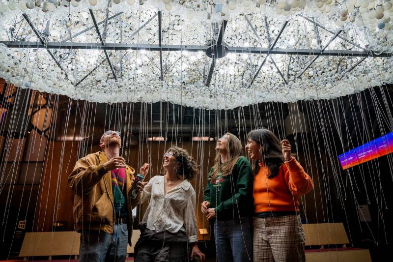 Four people smile up at lit sculpture made of light bulbs with chains dangling around them