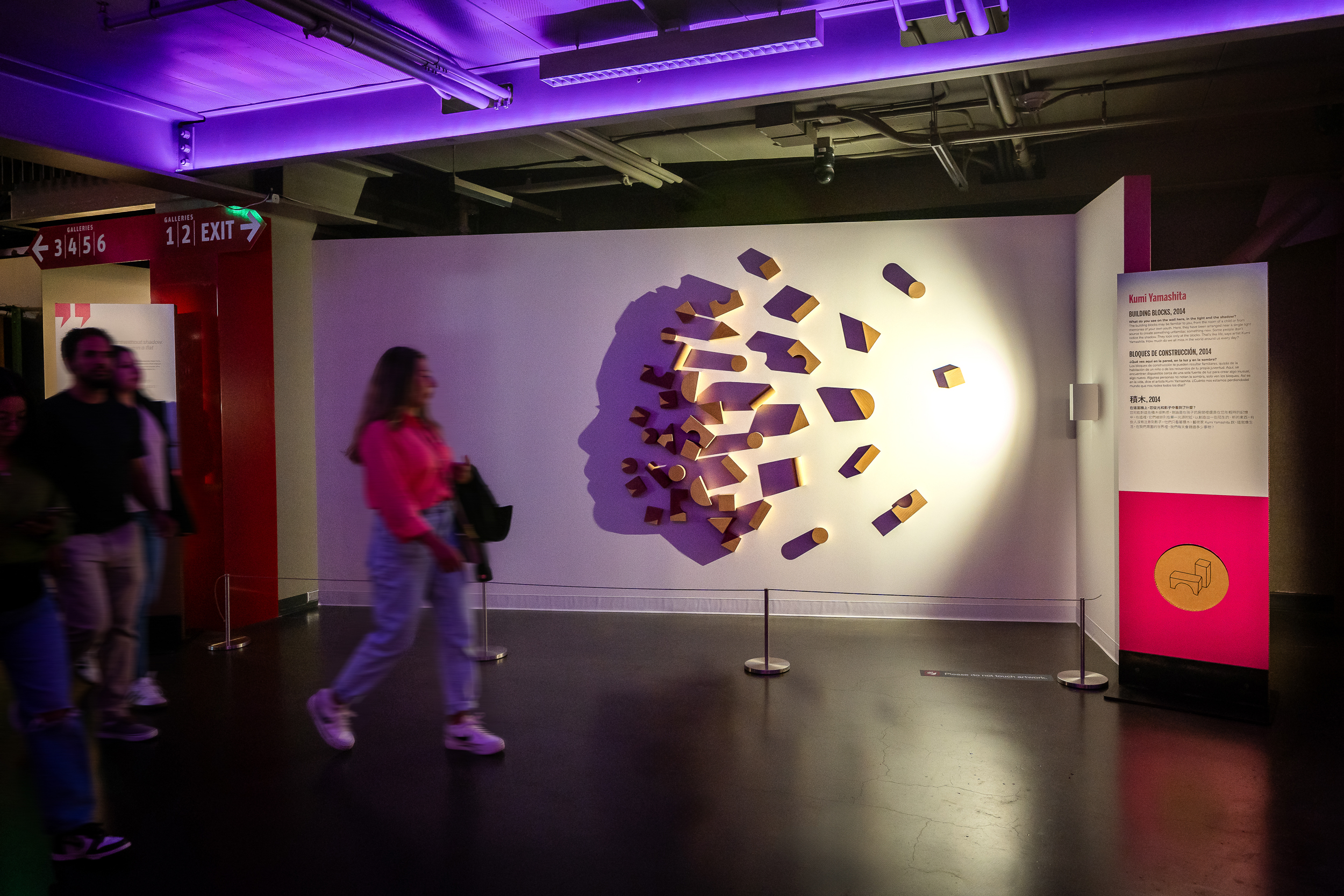 People walk past a wall display of lit wooden blocks that create a shadow silhouette