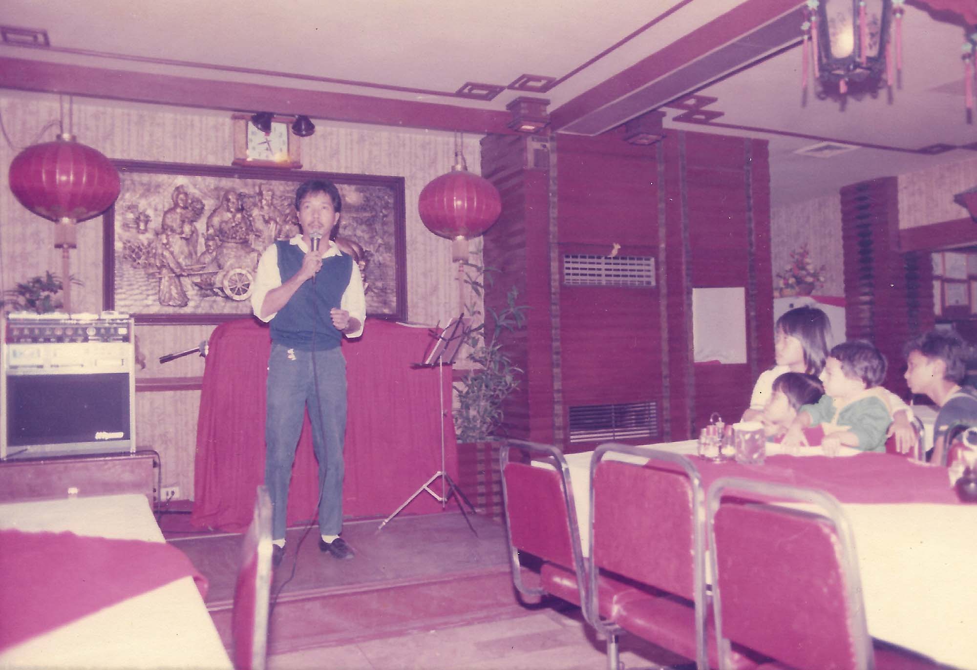 Vintage photo of a man singing karaoke at a Filipino restaurant in the 1980s. A group of children seated at a table look on.